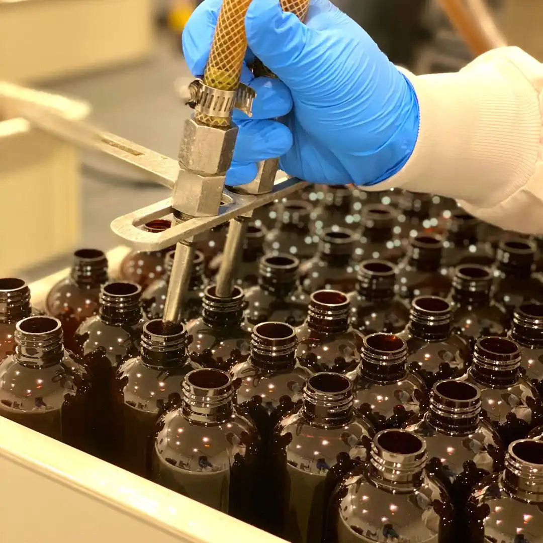 Gloved hand filling dark glass bottles using a dispensing nozzle.